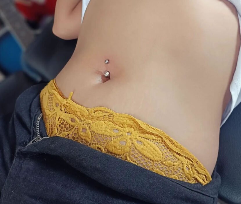 Close-up of a navel piercing with two studs, featuring a person wearing a white top, dark pants, and yellow lace-trimmed underwear.