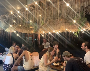 A group of people dining together at rustic wooden tables under warm string lights in an open-air setting.