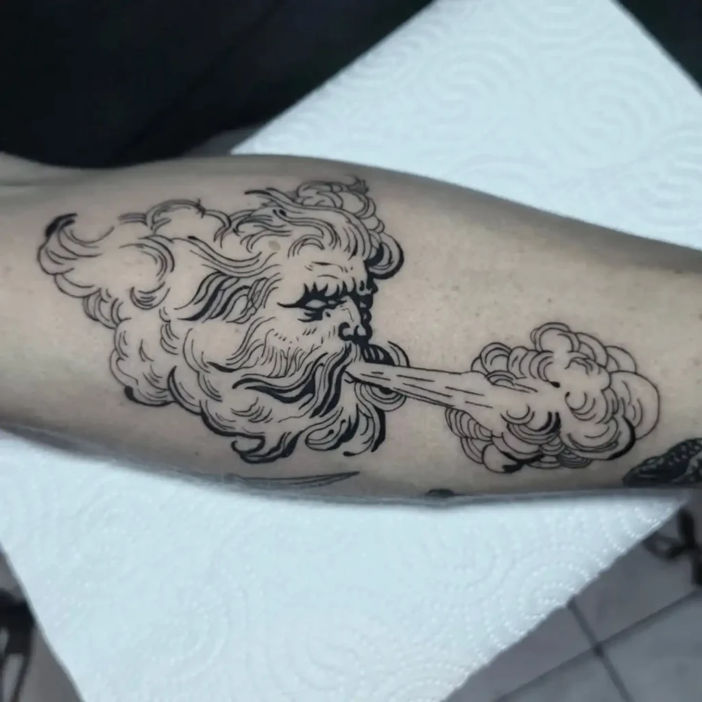 Close-up of a tattoo on an arm depicting a bearded face blowing wind, with swirling clouds extending across the skin.