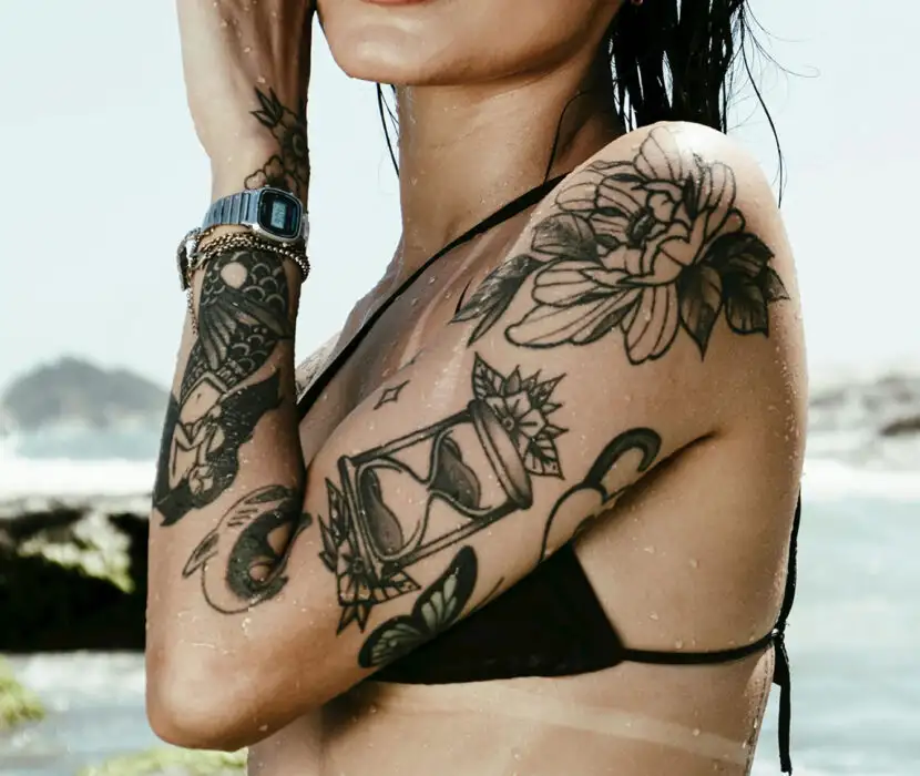 Person with tattoos, black bikini top, wristwatch, and bracelets, standing near ocean waves and rocks.