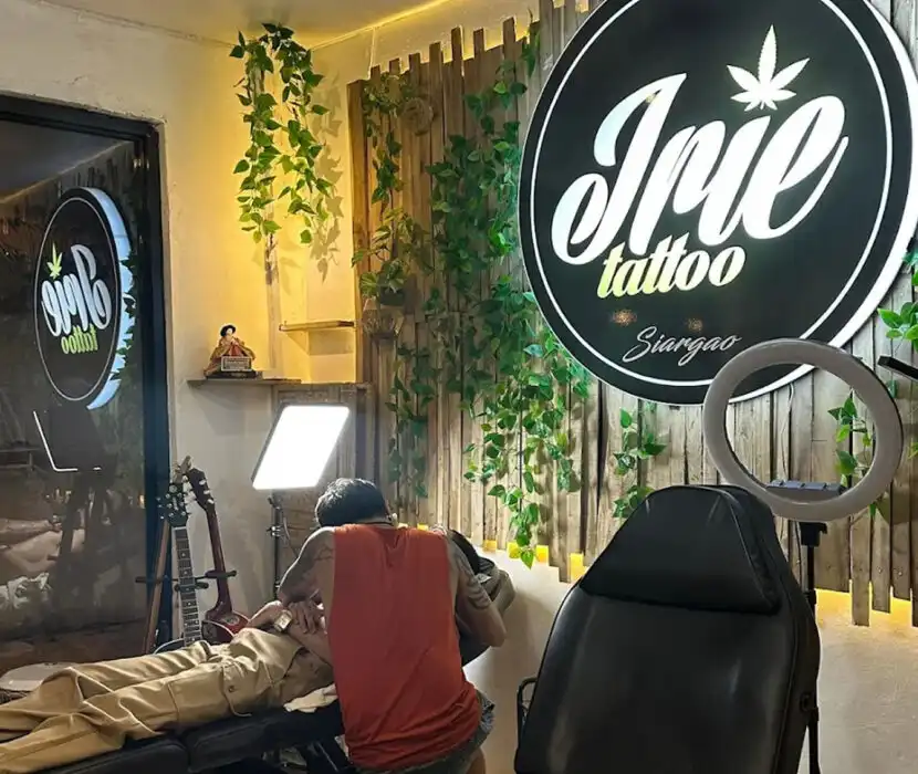 Interior of Irie Tattoo studio in Siargao, showing a tattoo artist working on a client lying on a table, with a guitar, ring light, and green plants against a wooden wall.